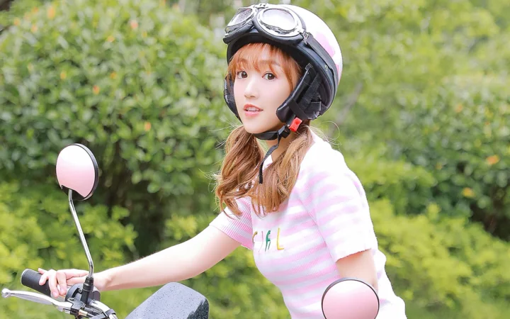 一名戴着头盔的女性骑在踏板车上，身后是绿色树丛。
