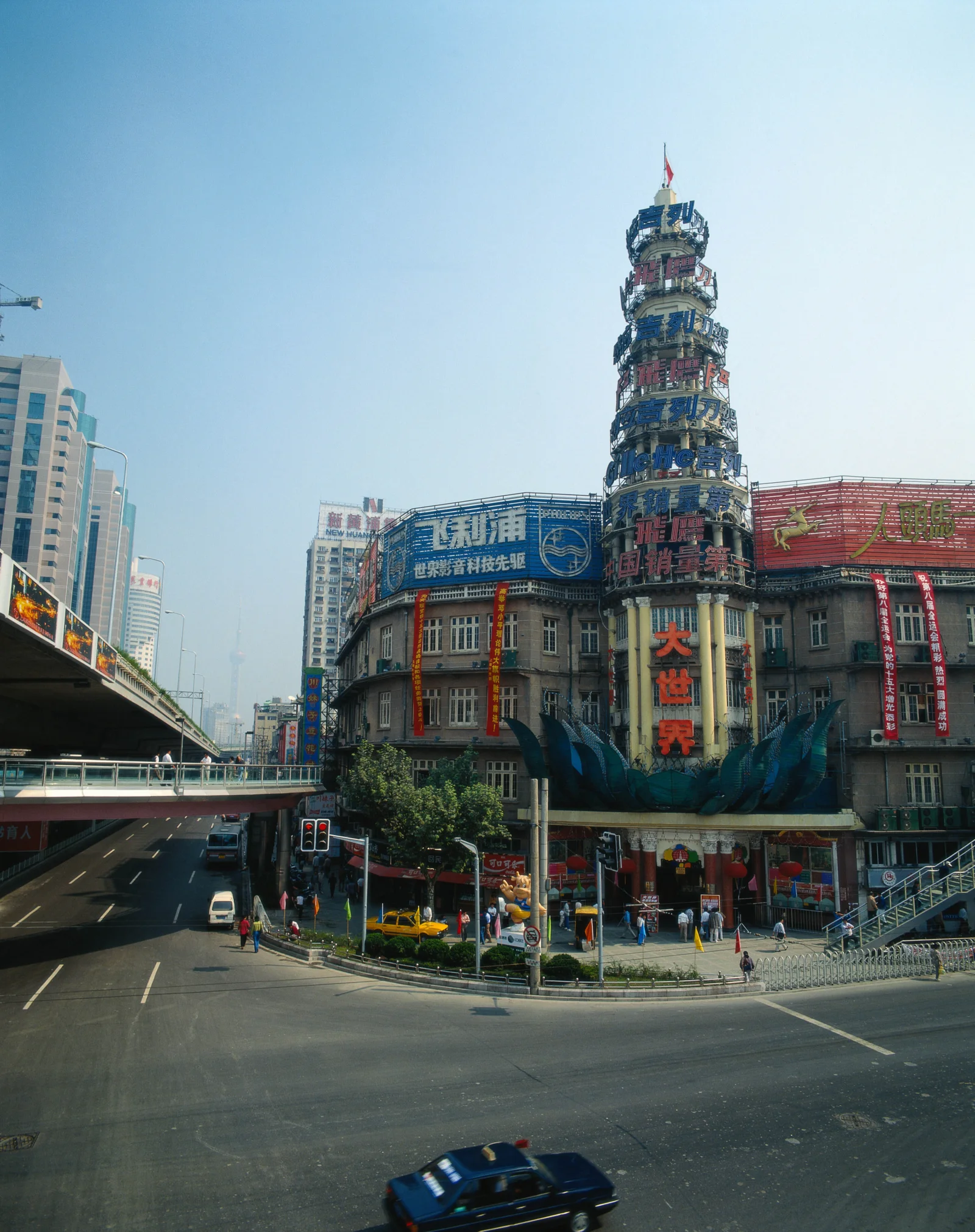 蓝天下的城市街道旁矗立着一座多层商业建筑，前方可见高架道路、路口和少量车辆。