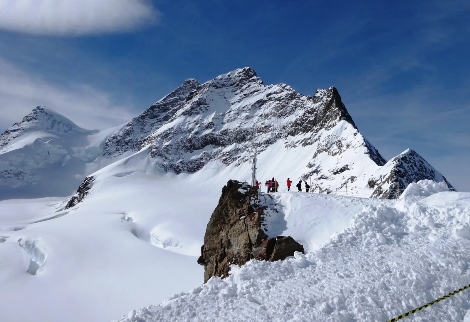 白雪覆盖的险峻山峰在蓝天下延伸，山脊上可见几名远处的登山者。