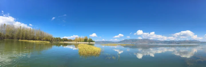 湛蓝天空下的湖面倒映着远山、白云与岸边芦苇，构成开阔宁静的自然全景。