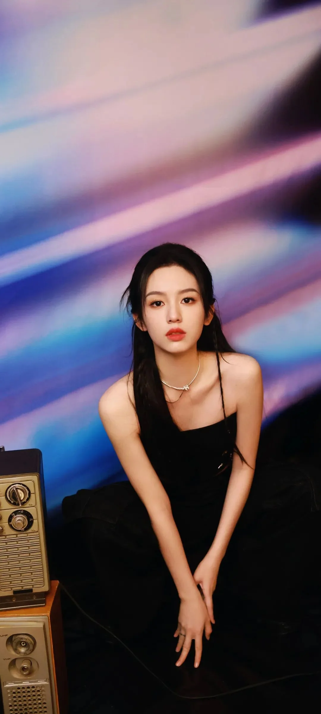 一名身穿黑色服裝的女性坐在彩色光影背景前。