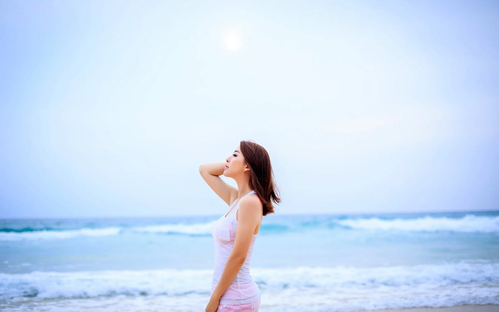 一名女子站在海边，身后是明亮天空、海浪与阳光。