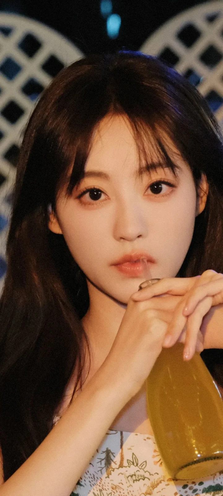 一位长发女性在暖色光线下以双手托腮，背景带有几何图案。