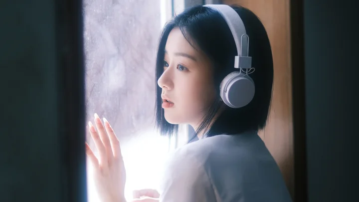 헤드폰을 쓴 여성이 창가에 서 있고 창밖에서 부드러운 빛이 들어온다.
