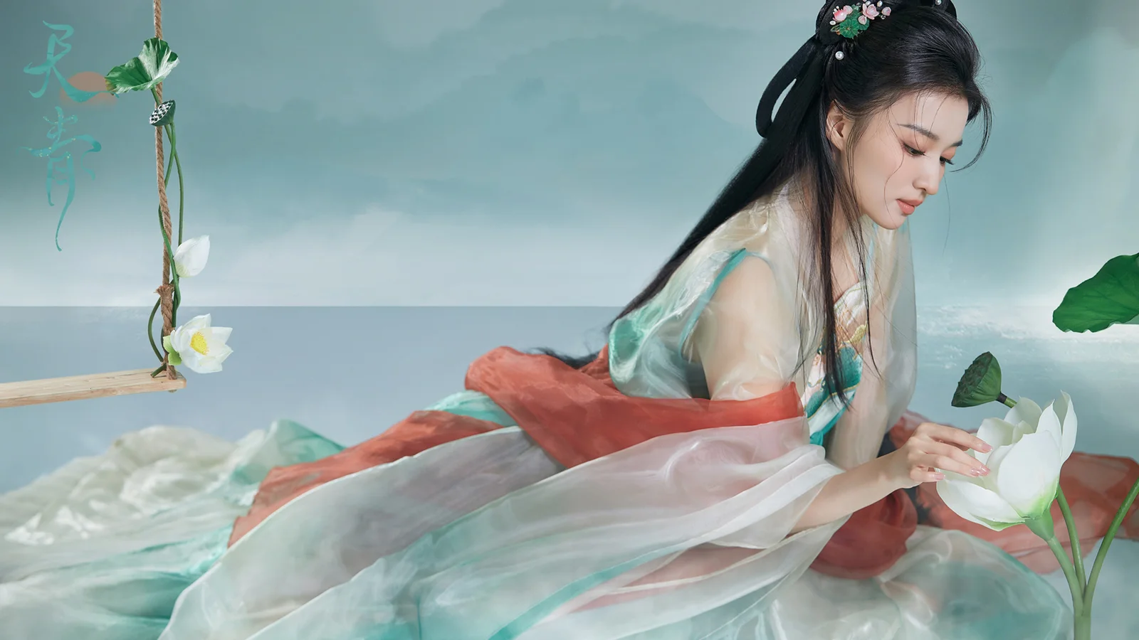 一名身穿浅色传统服饰的成年女性坐在荷花旁，画面以蓝绿色调为主。
