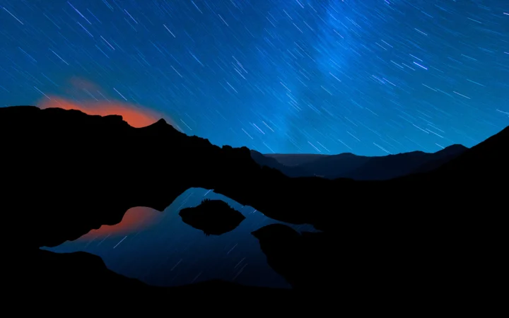 夜空中的星轨划过群山与湖泊上方，远处山脉和湖面呈现深色剪影。