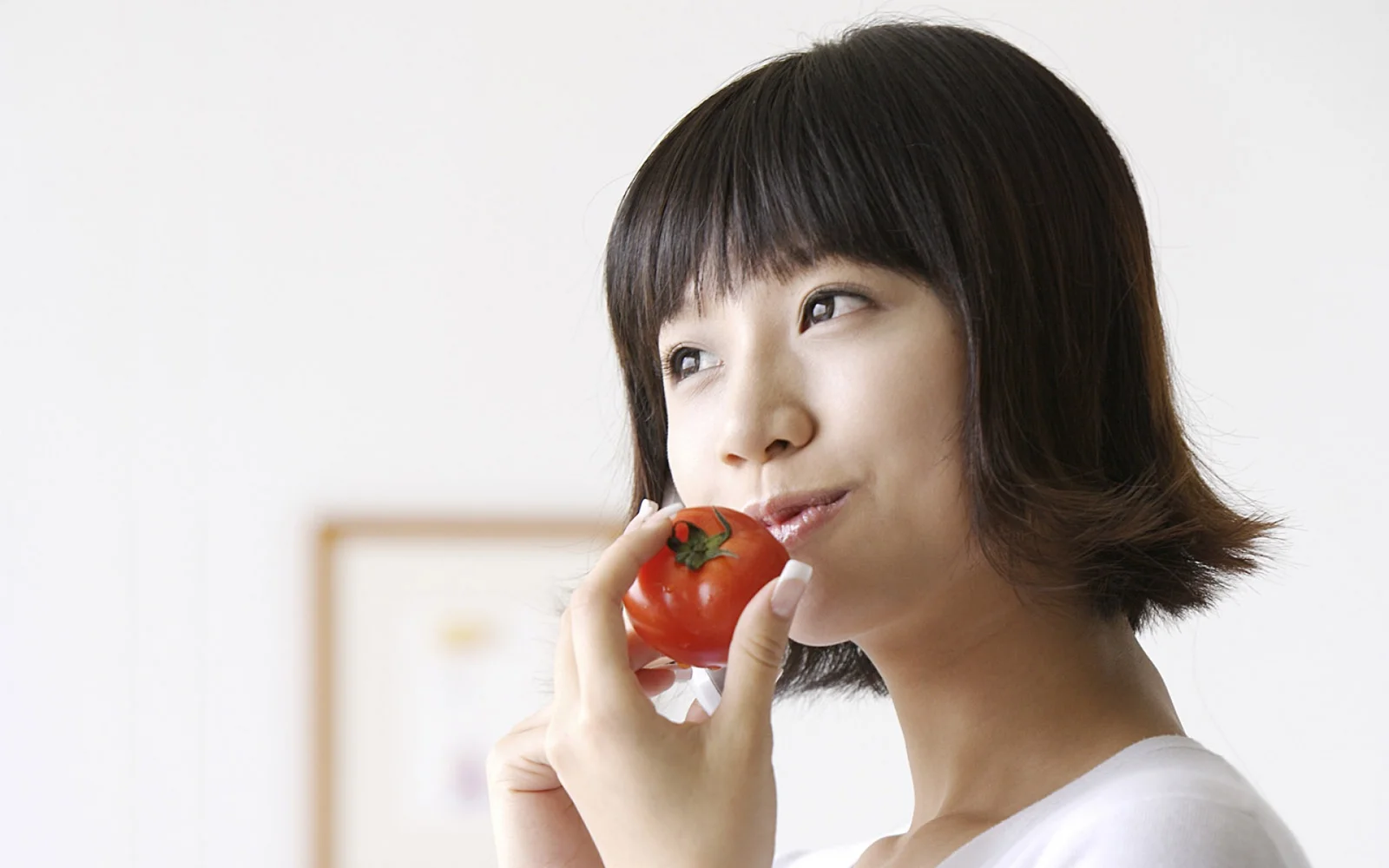 明るくシンプルな背景で、女性がトマトを口元に持っているポートレート。