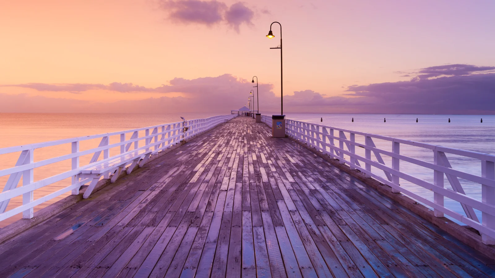 一座木質棧橋延伸向平靜的海面，沐浴在粉紫色的夕陽餘暉中。