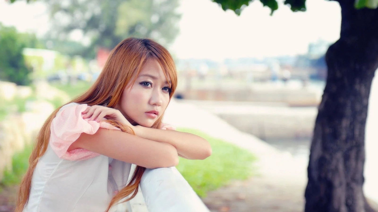 공원 나무 옆에 앉은 여성이 무릎 위에 팔을 올리고 앞을 바라보고 있습니다.