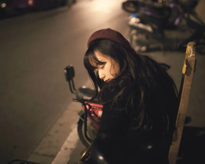 一名长发女子戴着棕色帽子坐在夜晚街边的自行车旁。