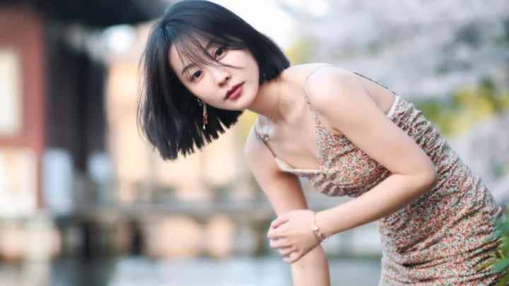 꽃무늬 원피스를 입은 단발머리 여성이 야외 물가에서 몸을 숙인 채 카메라를 바라보고 있습니다.