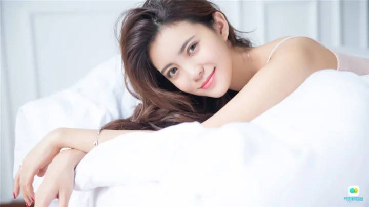 흰 침구 위에 누워 카메라를 향해 미소 짓는 성인 여성.