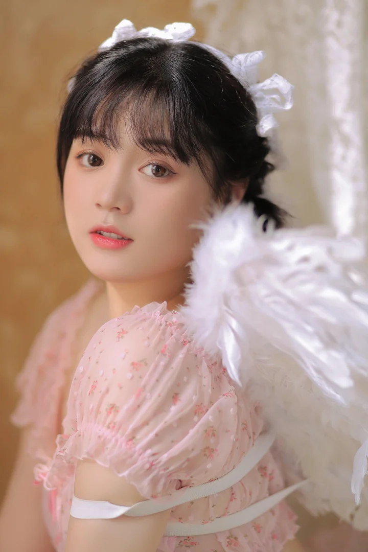 ピンクの柄入りドレスと白い髪飾りを身に着け、大きな白い羽根の翼を添えた女性の近距離ポートレート。
