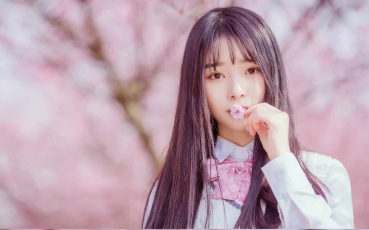 一名长发女子手持小花站在粉色樱花背景前。