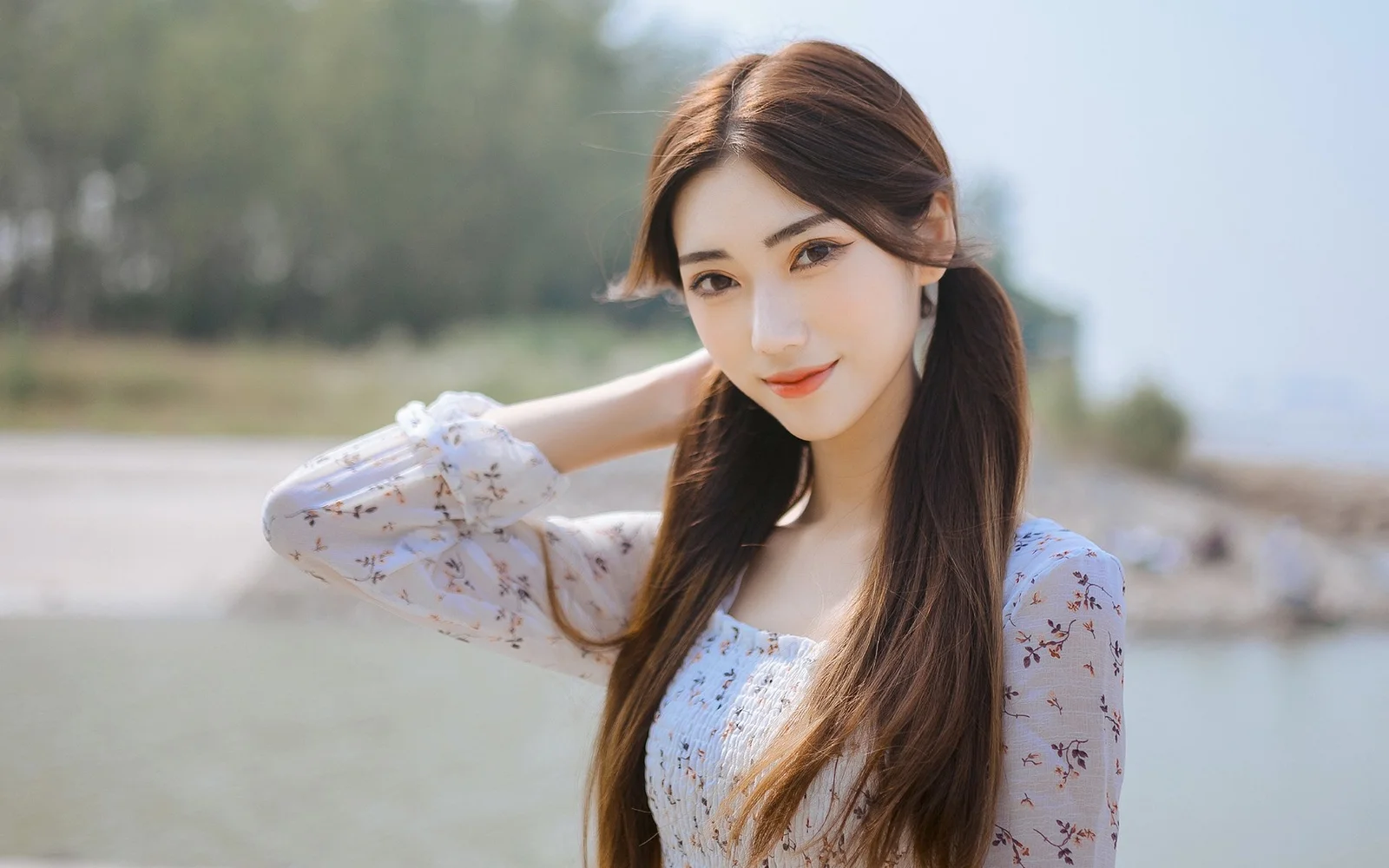 긴 머리의 여성이 밝은 꽃무늬 상의를 입고 야외 물가에서 카메라를 향해 미소 짓고 있다.
