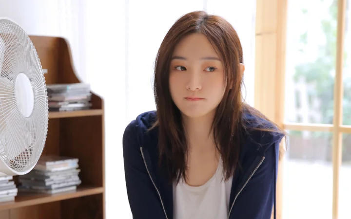 一名成年女性坐在明亮的室内，旁边可见电风扇和书架。