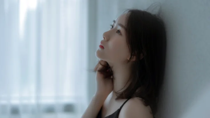 성인 여성이 벽에 기대어 창가의 부드러운 빛 속에서 옆모습을 보이고 있습니다.