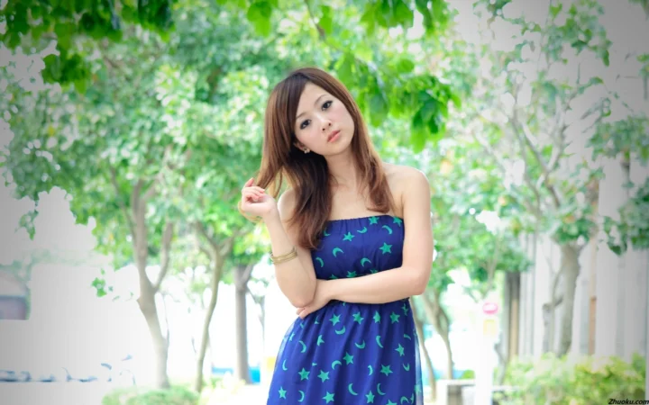 파란색 무늬의 끈 없는 원피스를 입은 여성이 푸른 나무로 둘러싸인 야외 길가에 서 있습니다.
