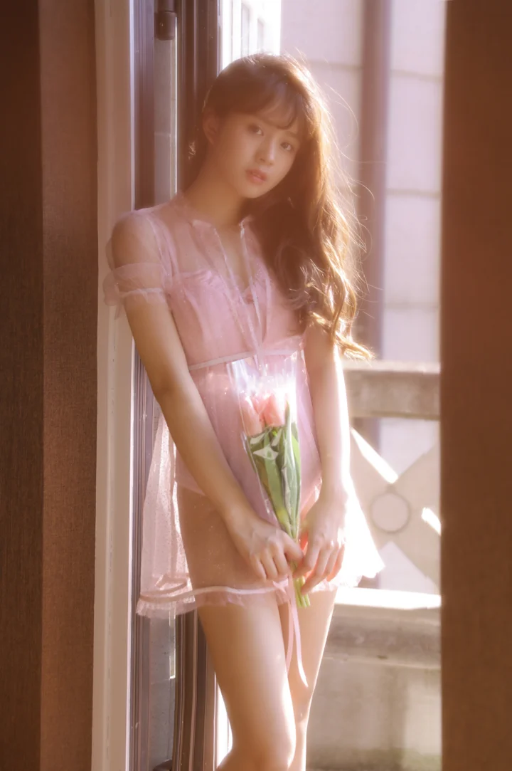 ピンクのワンピースを着た女性が花を手に窓辺に立ち、柔らかな日差しに包まれています。