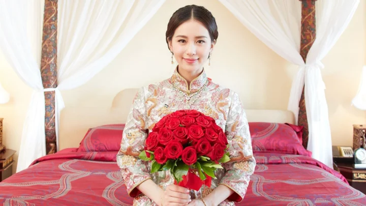 一名身穿传统服饰的女子坐在床前，手捧一束红玫瑰。