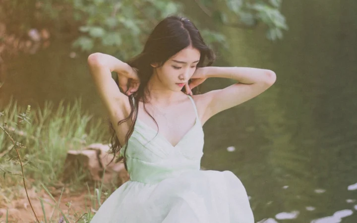 一名身穿白裙的成年女子坐在林木环绕的水边。