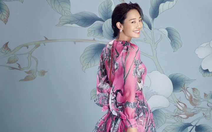一名身穿粉色花卉图案服装的成年女性站在淡蓝色花卉背景前微笑回望。