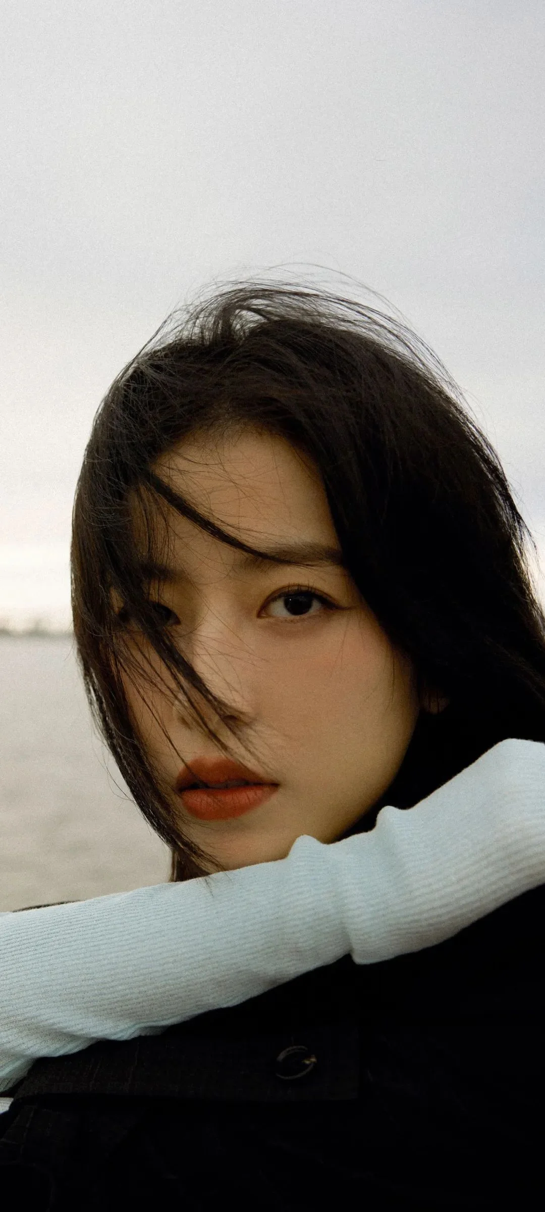 一名女性在海边近距离面对镜头，黑发被风吹拂，背景可见浅色天空与海面。