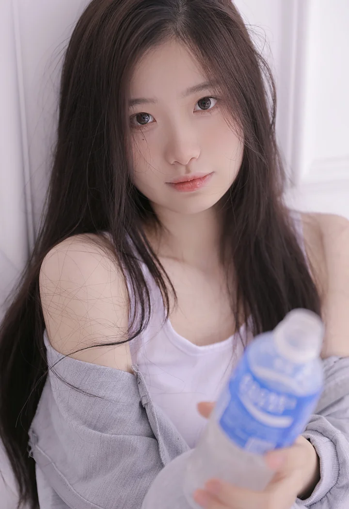 一名长发女性身穿白色上衣和浅灰色外套，在室内手持蓝色水瓶面对镜头。