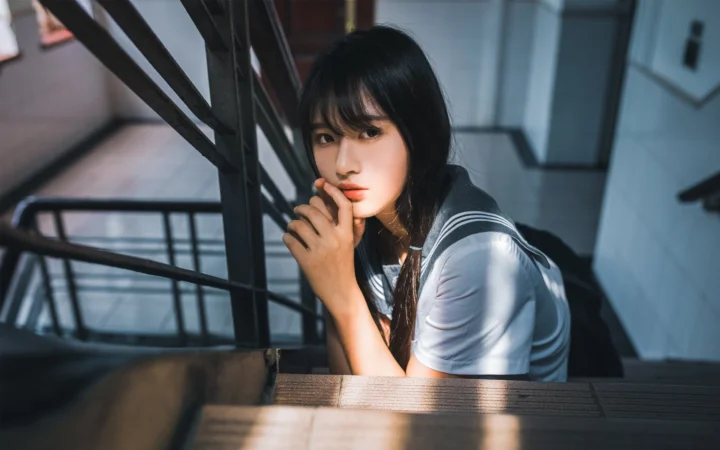 一名成年女性站在室内楼梯间的栏杆旁，阳光照亮了她的侧脸和上衣。
