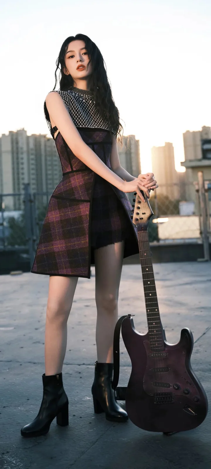 一名身穿深色格紋連身裙和短靴的女性站在城市建築間，手扶一把電吉他。