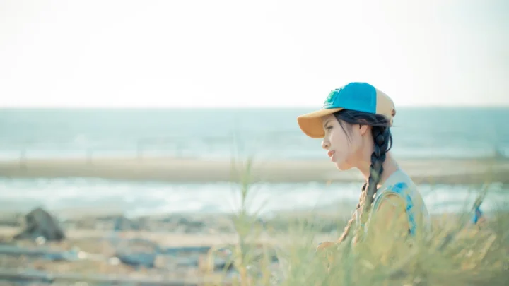 一名戴帽子的成年女性侧身坐在海边草丛中，远处是海面与沙滩。