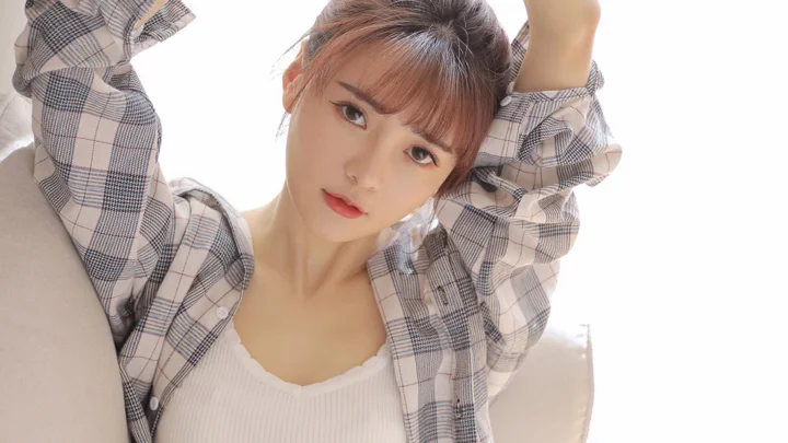 체크무늬 셔츠와 흰색 캐미솔을 입은 성인 여성이 팔을 올리고 카메라를 향해 포즈를 취하고 있다.