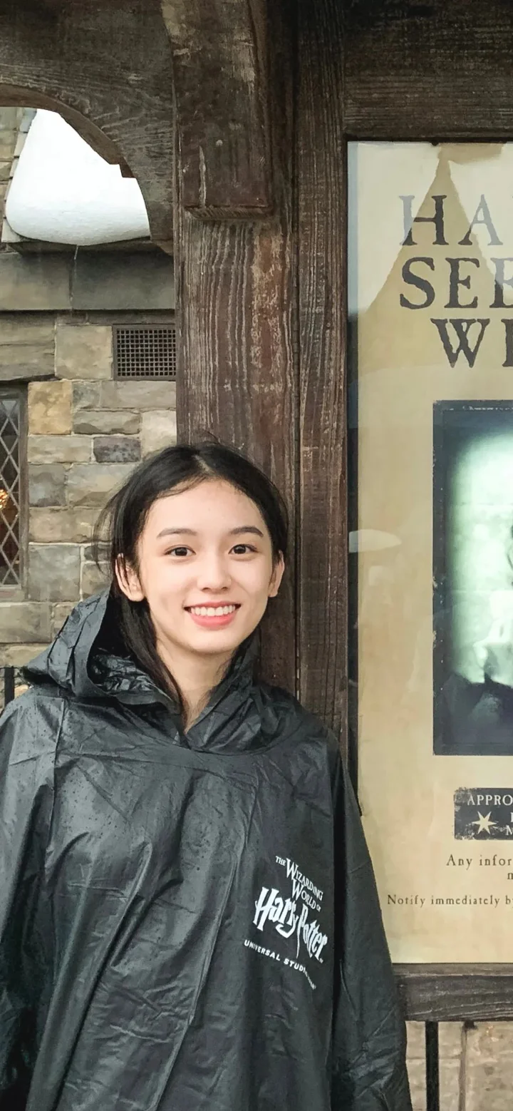 一位面带微笑的成年女性穿着黑色雨衣，站在木质建筑旁。