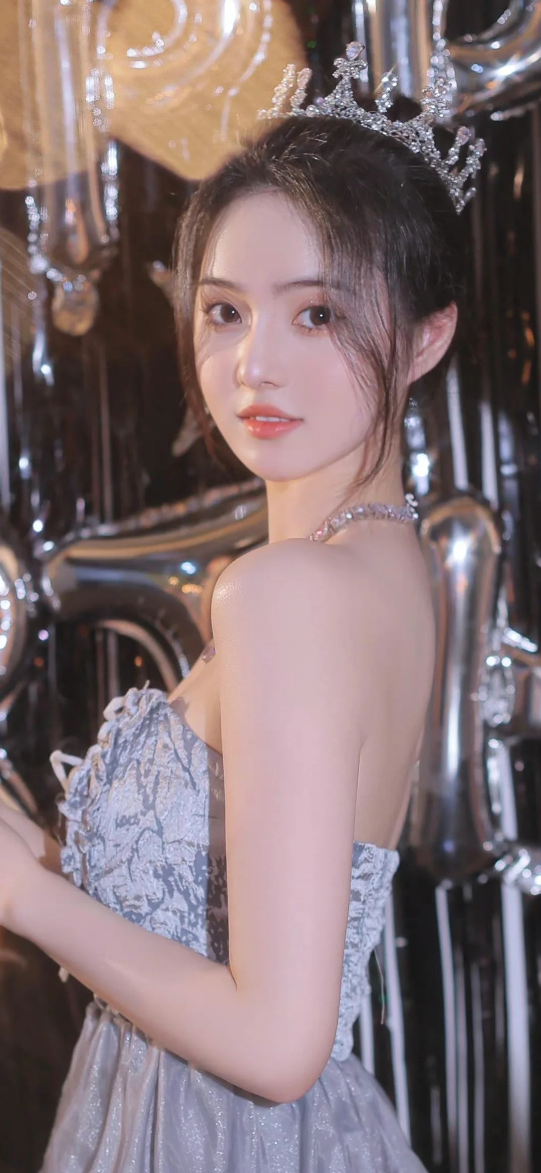 一位戴著皇冠、身穿銀色禮服的女性在金屬裝飾背景前回眸。