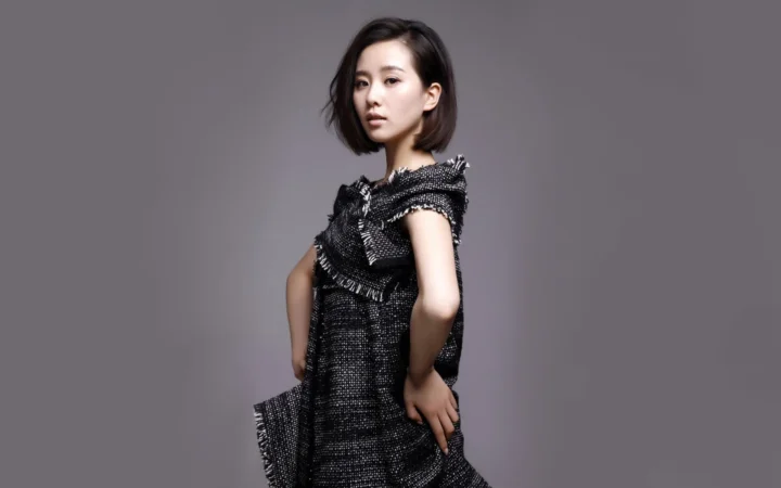 짧은 머리의 여성이 검은색 드레스를 입고 회색 배경 앞에 서 있습니다.