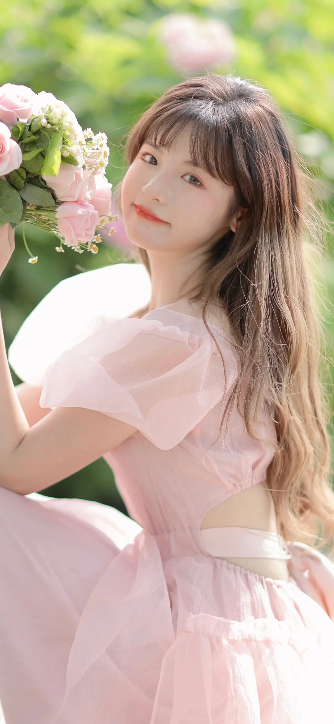 一名长发女子身穿粉色连衣裙，在户外手捧鲜花。