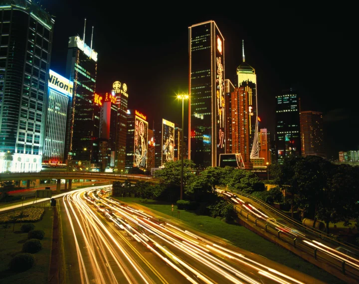 夜间高楼与霓虹广告牌映照在道路上，车流留下明亮的光轨。