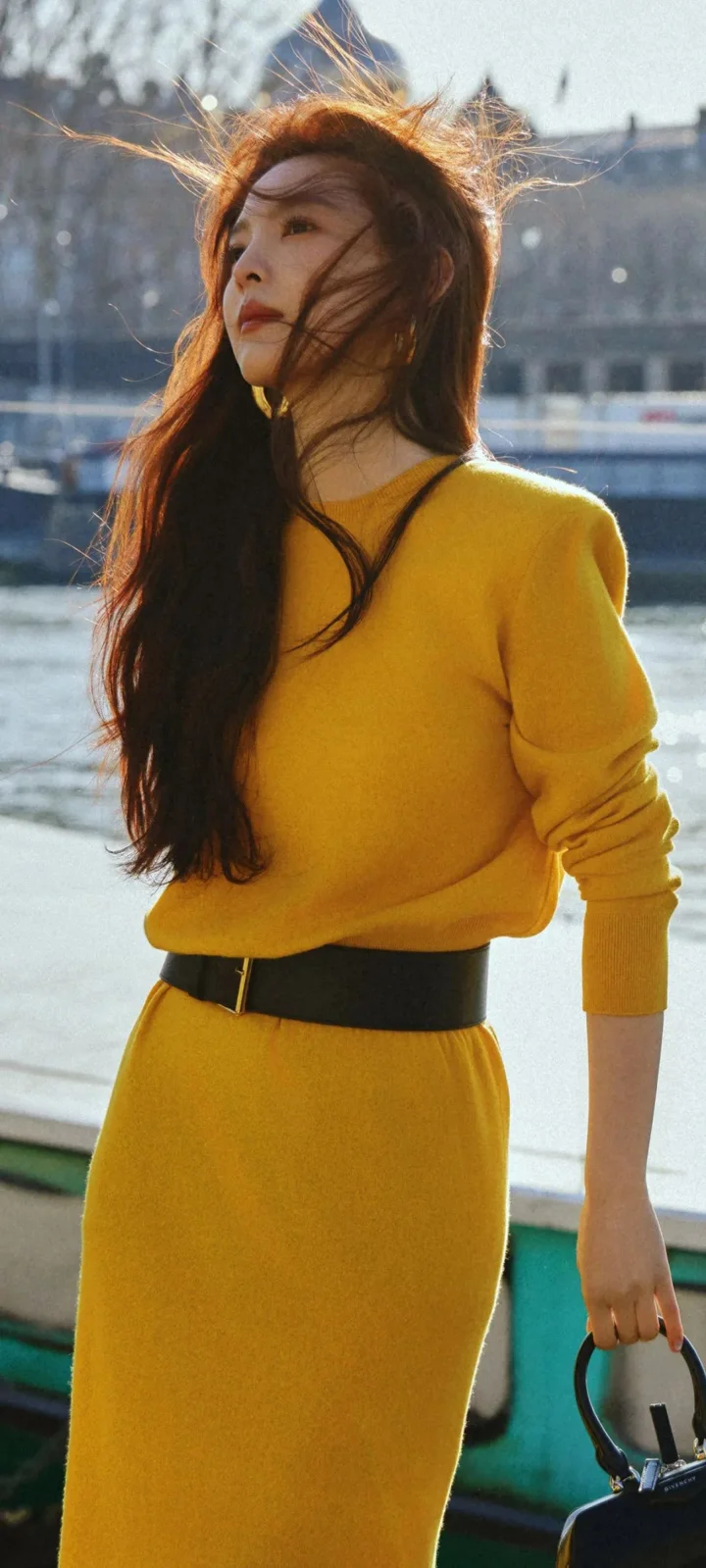 긴 머리의 여성이 노란색 상의와 치마를 입고 햇빛이 비치는 야외 물가에 서 있다.