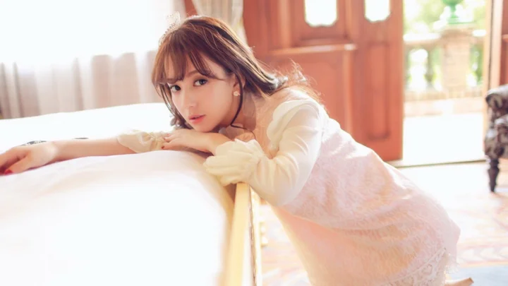 성인 여성이 침대 가장자리에 기대어 있고 따뜻한 창가의 빛이 실내를 비추고 있다.