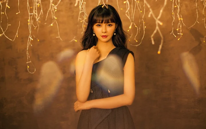 一名成年女性身穿深色无袖服装站在暖棕色背景前，头顶悬挂着细小灯饰。