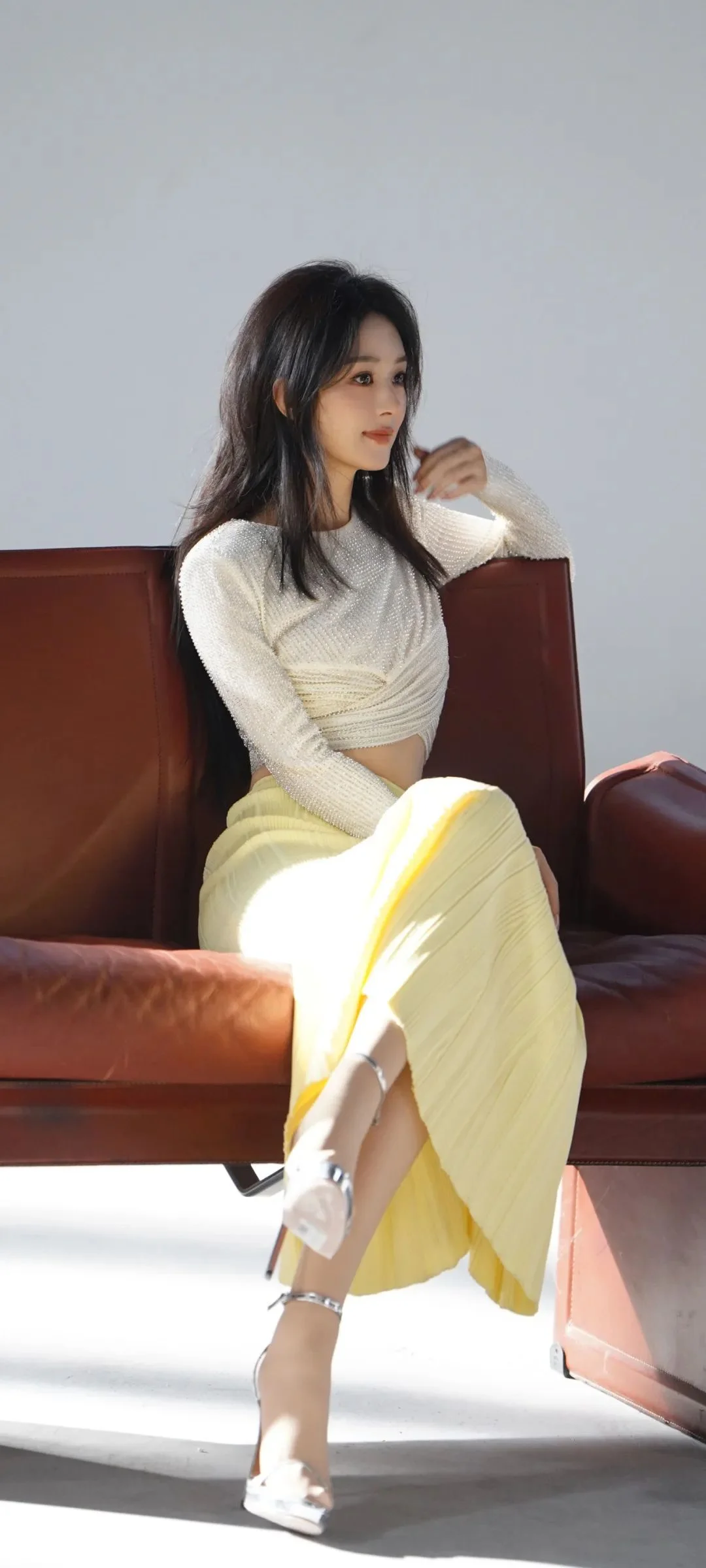 一名女子穿着白色上衣和浅黄色裙子，坐在棕色皮沙发上，阳光照亮了室内空间。