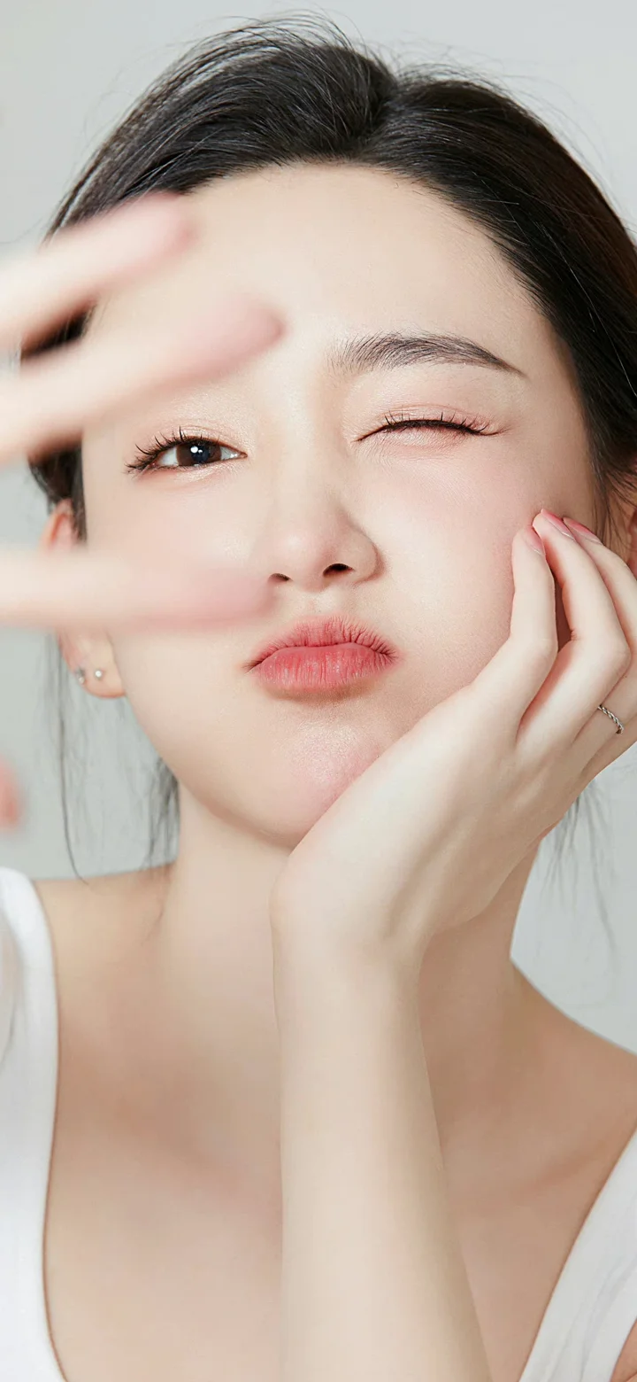一位女性近距离面对镜头眨眼并用手托着脸颊，背景简洁明亮。