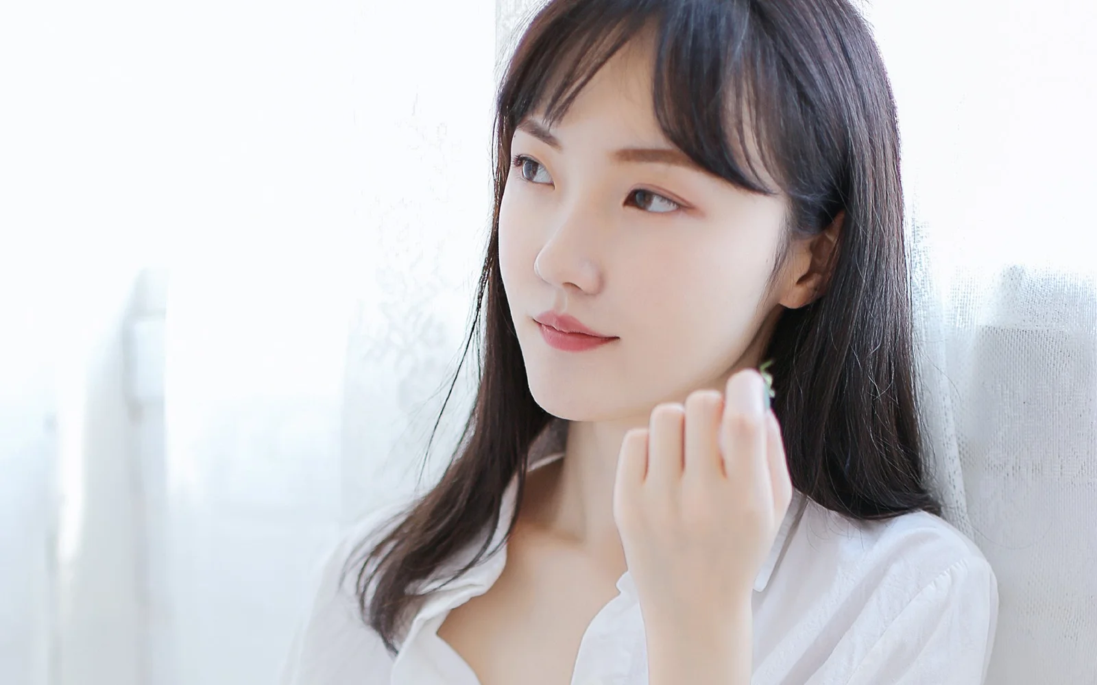 흰 셔츠를 입은 성인 여성이 밝은 창가에 서서 옆모습으로 먼 곳을 바라보고 있습니다.