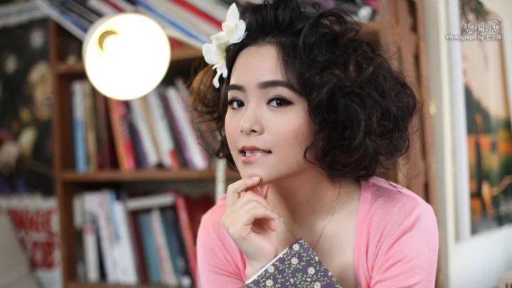 곱슬머리에 흰색 꽃 장식을 한 성인 여성이 책장 앞에서 카메라를 바라보고 있습니다.