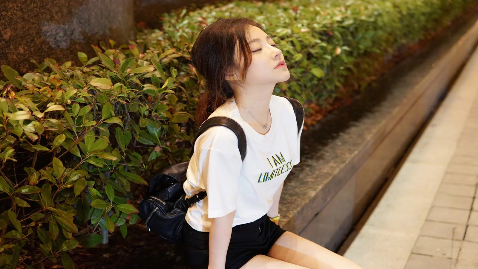女性が生け垣沿いの歩道脇に座り、目を閉じて自然光を浴びている。