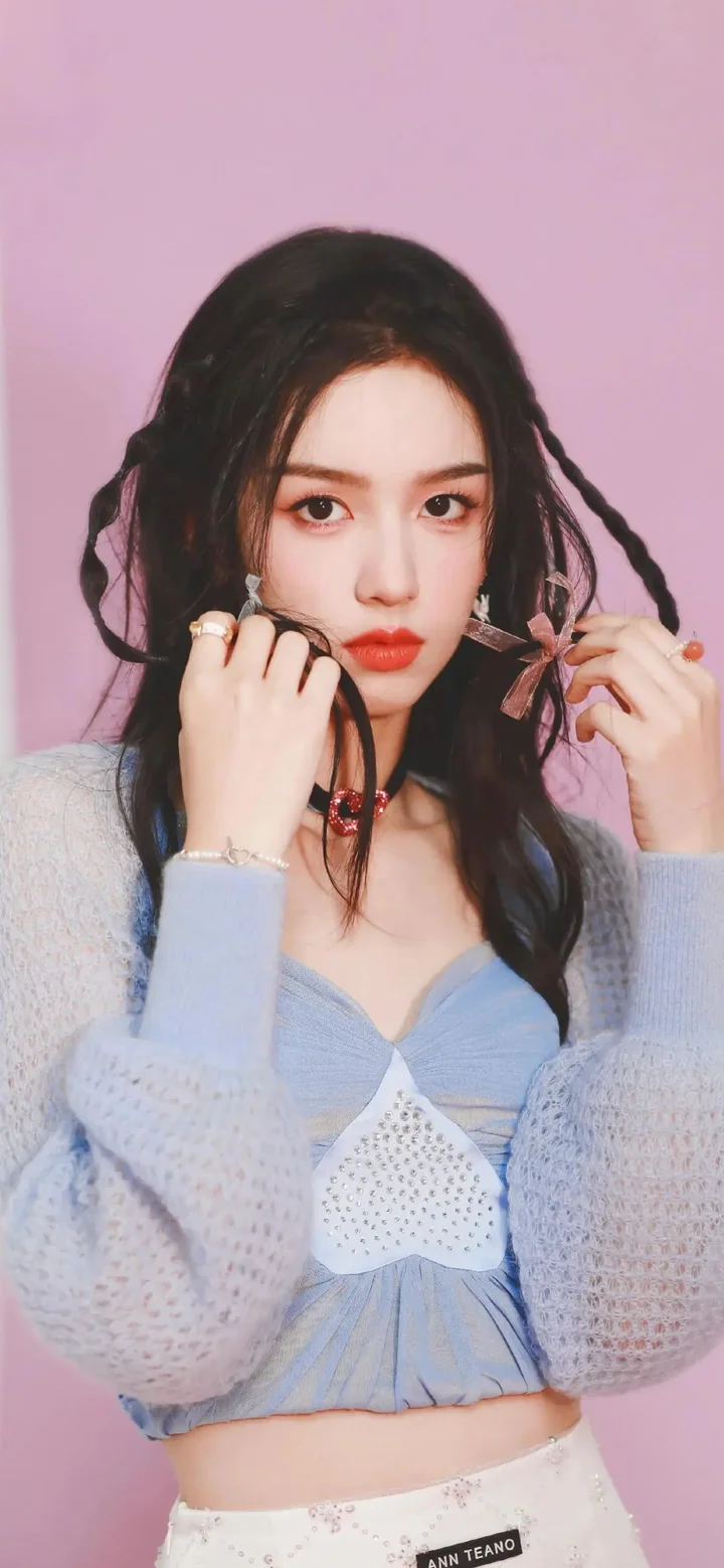 분홍색 배경 앞에서 연한 파란색 니트 차림의 검은 머리 여성이 머리를 정리하고 있습니다.