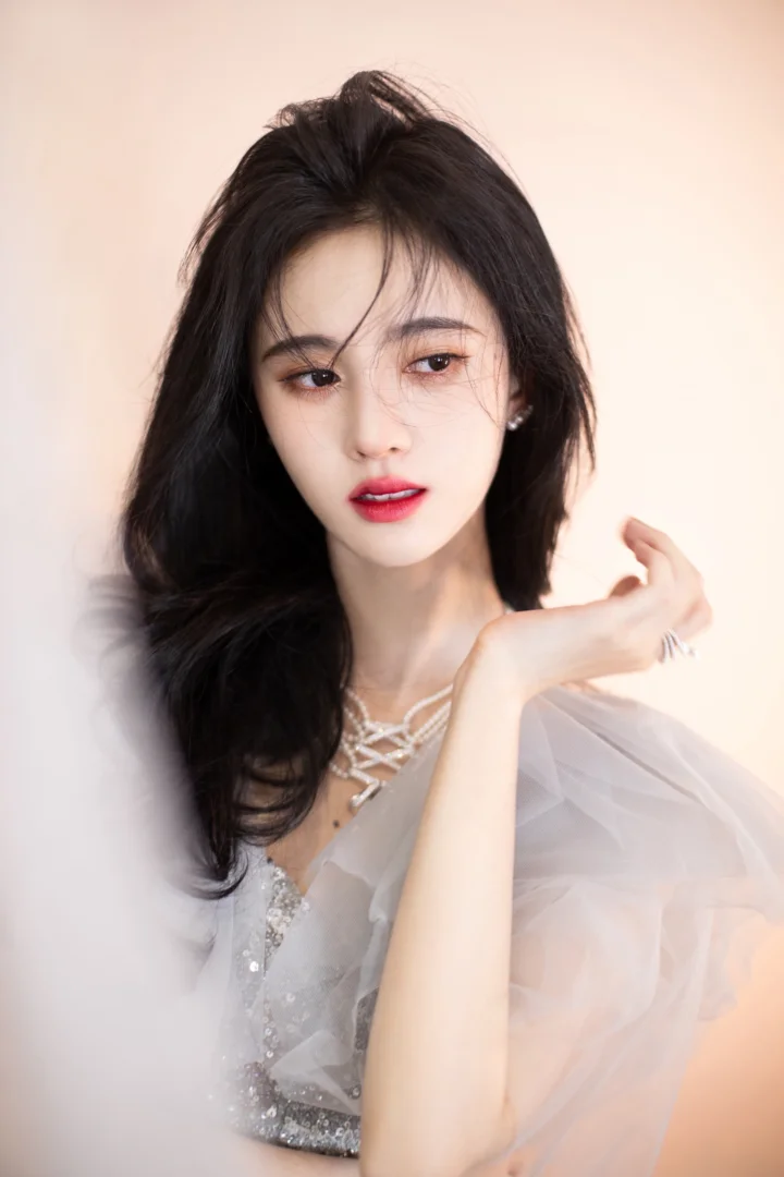 一位黑发女性身穿浅色薄纱礼服，佩戴项链，在柔和的浅色背景前摆出优雅姿势。