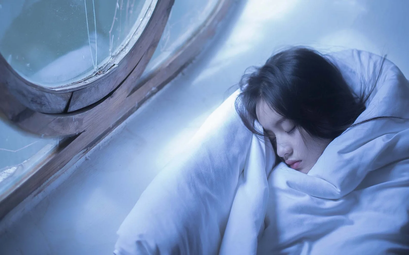 한 여성이 창가에서 흰 침구에 싸여 차가운 푸른빛 속에 잠들어 있습니다.