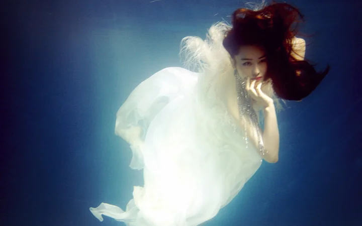白い流れるようなドレスを着た女性が青い水中で浮かんでいる。
