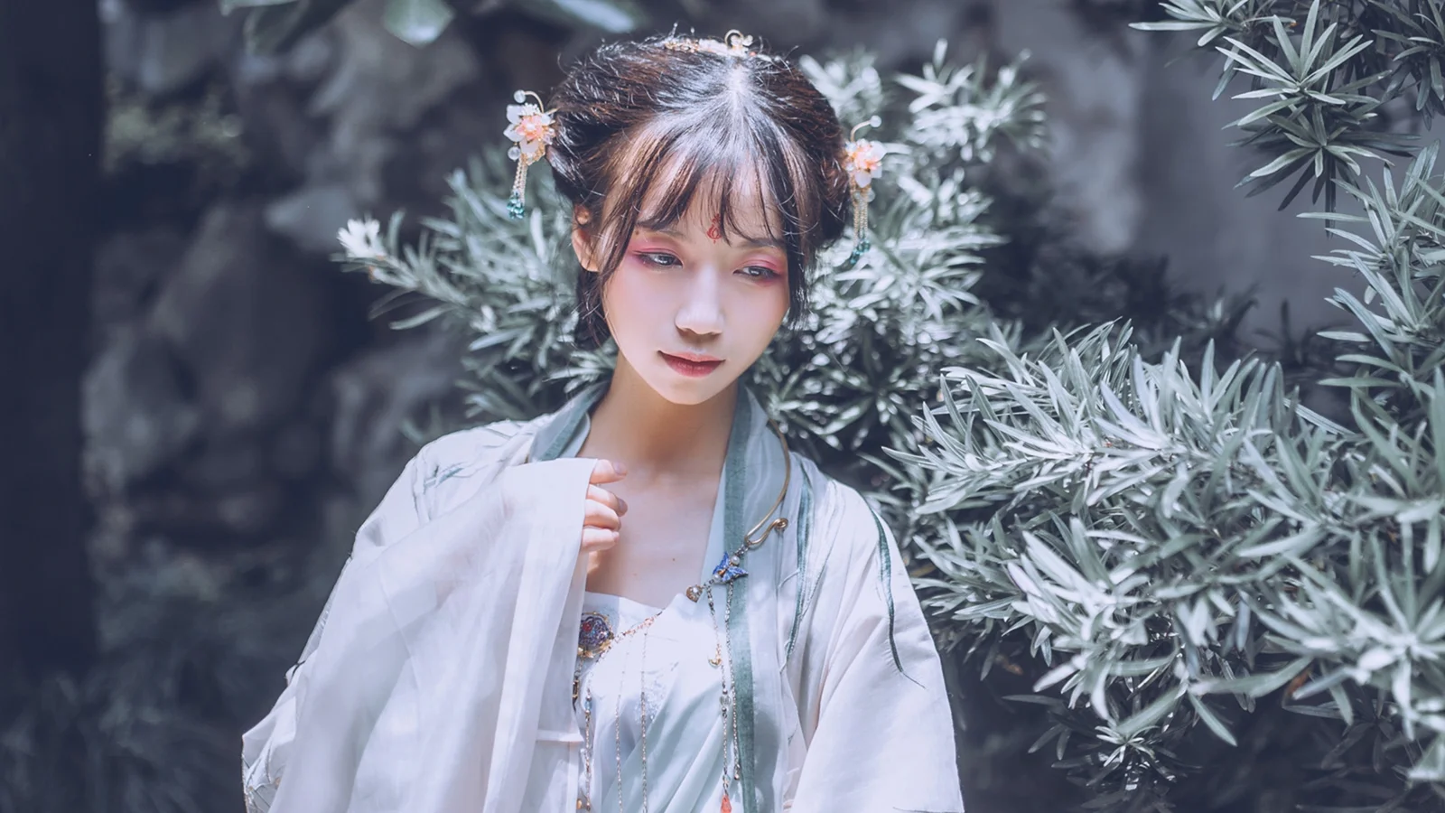 銀緑色の葉に囲まれた女性が、淡い色の衣装と髪飾りを身に着けて立っている。